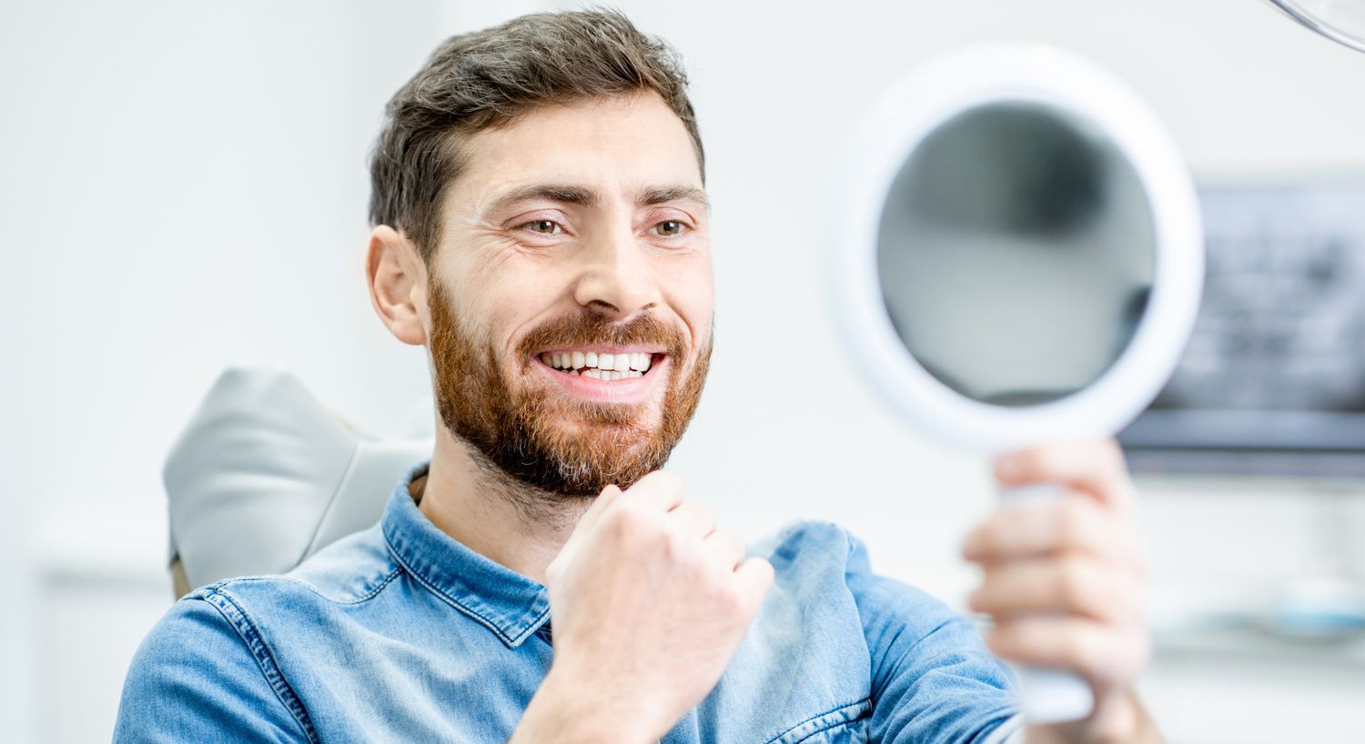 Smiling man examines teeth in handheld mirror.