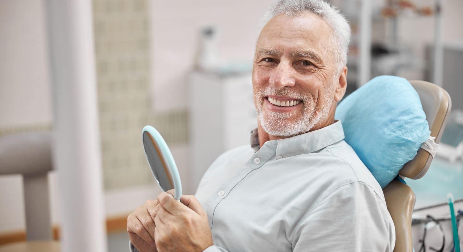 Smiling senior man at dental appointment holding mirror.